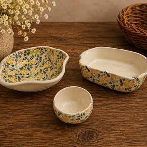 May include: A set of three ceramic dishes featuring a speckled cream base with a delicate floral pattern of yellow flowers and green leaves. The set includes one irregular oval bowl, one rectangular dish with rounded edges, and one small round bowl.