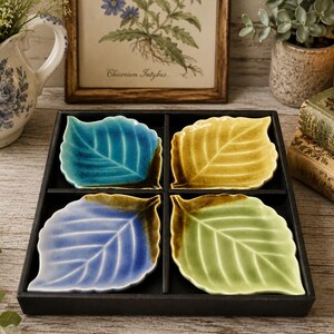 Può includere: Un set di quattro piattini in ceramica a forma di foglia disposti in un vassoio quadrato nero. Ogni piattino presenta un colore distinto, tra cui turchese, giallo, blu e verde chiaro, con accenti marrone scuro lungo i bordi e i gambi.