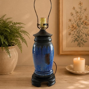 May include: A table lamp featuring a glossy, cylindrical ceramic base with a vibrant blue glaze that transitions into darker tones. The lamp includes a brass-colored socket and a metal harp, all resting on a sturdy, black, tiered circular base.