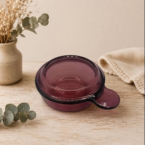 May include: A small, round, translucent purple glass dish with a matching lid and a single side handle. The dish features a smooth, polished surface and a circular design, providing a clear view of the interior through the deep purple glass.