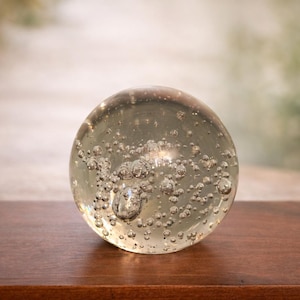 Puede incluir: Una esfera de vidrio transparente con numerosas burbujas de aire suspendidas en su interior. La esfera está colocada sobre una superficie de madera marrón oscuro, con un fondo borroso de tonos neutros. Esta bola de cristal decorativa podría usarse como pisapapeles o decoración del hogar.