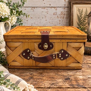 Op de afbeelding: Een rechthoekige mand van gevlochten hout met een scharnierend deksel, voorzien van een donkerbruin lederen handvat en een bijpassende lederen sluiting met een goudkleurig metalen slot. De mand heeft een natuurlijke lichtbruine afwerking.