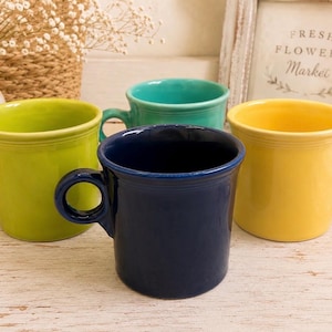 Puede incluir: Un conjunto de cuatro tazas de cerámica en colores sólidos que incluyen verde lima, turquesa, azul oscuro y amarillo brillante. Cada taza presenta un cuerpo cilíndrico con un asa redondeada y una textura anillada sutil en el borde superior.