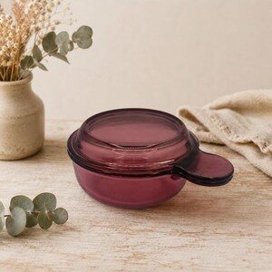 May include: A small, round, deep purple glass bowl with a matching lid and a single protruding handle on the side. The glass is translucent, allowing light to pass through the deep violet hue, and the lid features a slightly raised circular center.