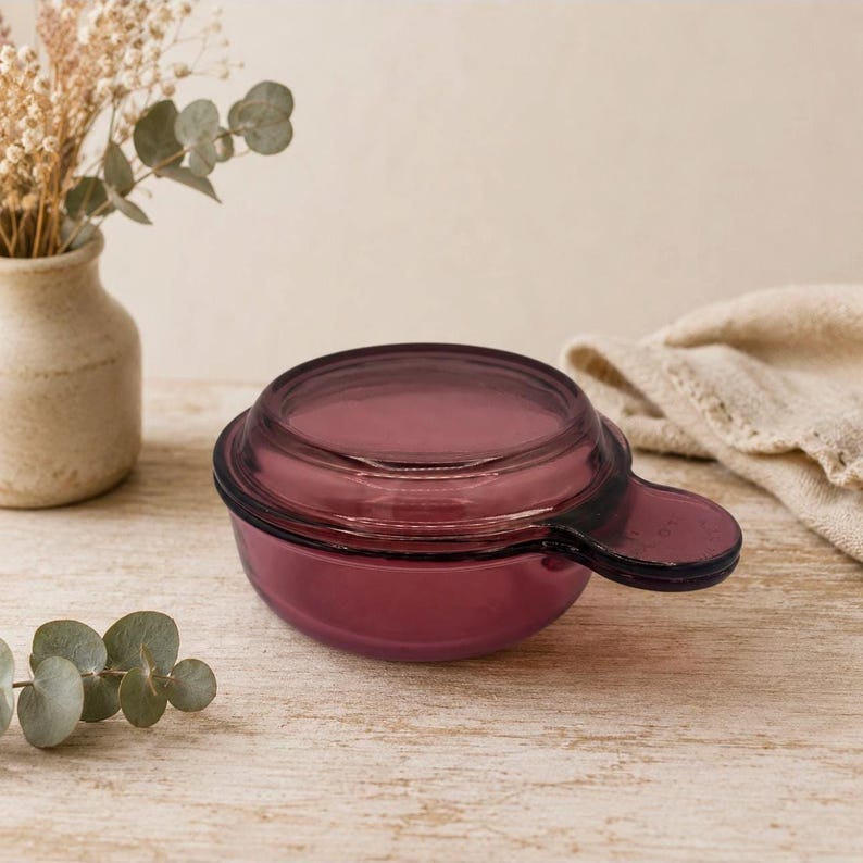 May include: A small, round, deep purple glass bowl with a matching lid and a single protruding handle on the side. The glass is translucent, allowing light to pass through the deep violet hue, and the lid features a slightly raised circular center.