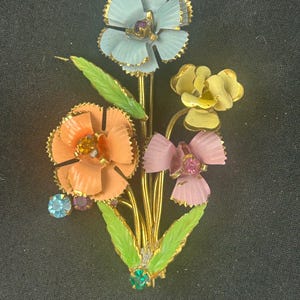 May include: A vintage floral brooch depicting a bouquet of flowers in pastel shades. The brooch features light blue, peach, yellow, and pink flowers with gold stems and green leaves. The flowers are embellished with jewel-toned accents.
