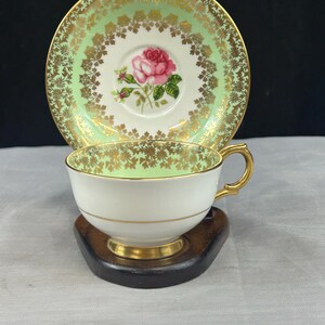 May include: A vintage teacup and saucer set. The cup and saucer have a white base with a light green and gold floral design. A pink rose is in the centre of the saucer. The cup has a gold handle and trim. The set is on a dark wooden stand.