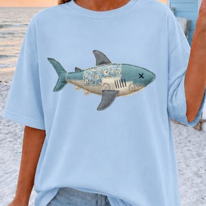 Puede incluir: Una camiseta azul claro con un gráfico central de un tiburón. El diseño del tiburón está compuesto por varios patrones de patchwork, incluyendo estampados florales y letras de estilo vintage, con un contorno cosido y una 'x' por ojo.