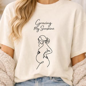 Puede incluir: Camiseta color crema con el texto "Growing My Sunshine" y un dibujo lineal negro de una persona embarazada. La camiseta tiene mangas cortas y un corte holgado.