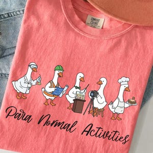 Puede incluir: Una camiseta de color coral con un gráfico de cinco gansos vestidos con diversas profesiones, incluyendo un científico, un obrero, un presentador, un fotógrafo y un chef. El texto "Para Normal Activities" está impreso en letra negra debajo.