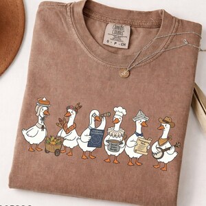 Könnte beinhalten: Ein braunes Kurzarm-T-Shirt mit einer Grafik von sechs weißen Gänsen in verschiedenen Outfits, darunter eine mit einem Tortenhut, eine mit einem Teleskop, eine mit einer Kochmütze und eine mit einem Banjo. Das Shirt hat ein Comfort Colors Markenetikett.