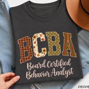 Könnte beinhalten: Ein dunkelgraues T-Shirt mit Rundhalsausschnitt und den Buchstaben BCBA im Patchwork-Stil mit braunem Karomuster, Sonnenblumen- und Blattmotiven, olivgrünen Punkten und Streifen. Darunter steht der Text Board Certified Behavior Analyst in Weiß.