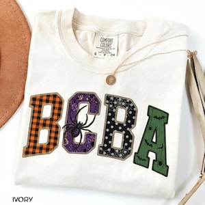 Könnte beinhalten: Ein elfenbeinfarbenes T-Shirt mit den Buchstaben BOBA in College-Schrift. Das B ist orange-schwarz kariert, das O ist lila mit einer Spinne, das B ist schwarz mit weißen Punkten und Spinnennetzen, und das A ist grün mit kleinen schwarzen Fledermäusen.
