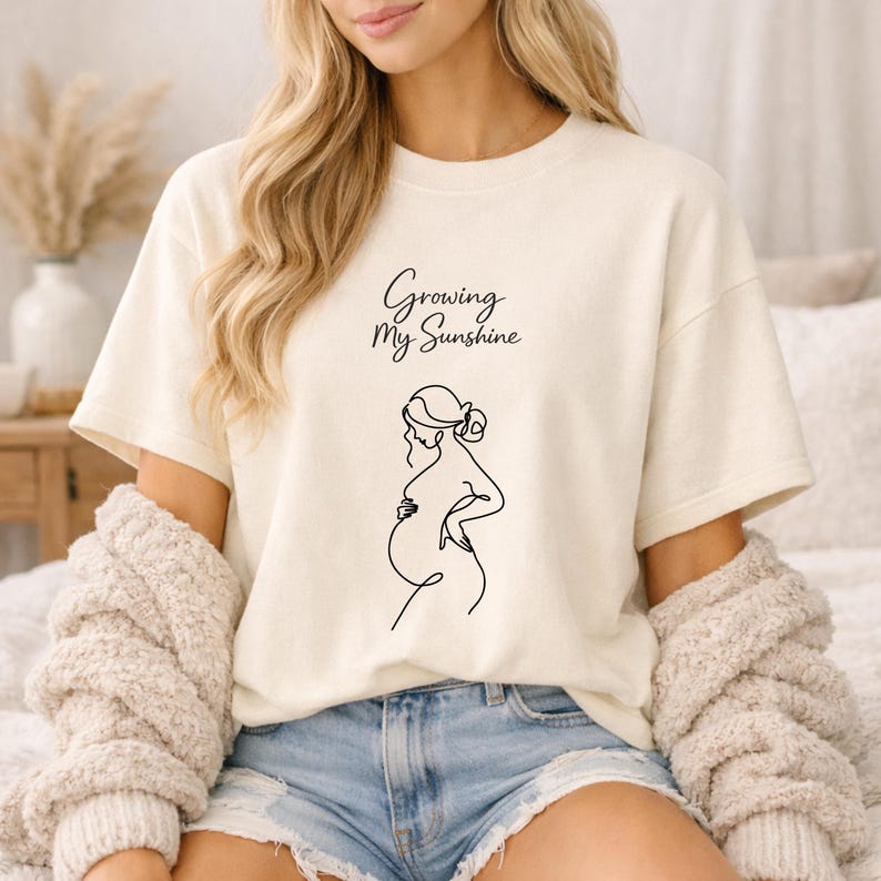 Puede incluir: Camiseta color crema con el texto "Growing My Sunshine" y un dibujo lineal negro de una persona embarazada. La camiseta tiene mangas cortas y un corte holgado.