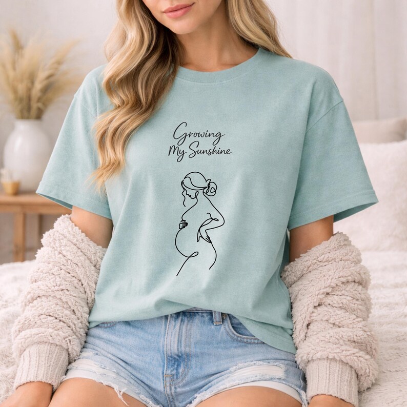 Puede incluir: Camiseta azul claro con el texto "Growing My Sunshine" y un dibujo lineal negro de una persona embarazada. La camiseta tiene mangas cortas y un corte holgado. La persona lleva pantalones cortos vaqueros y un su&eacute;ter color crema.