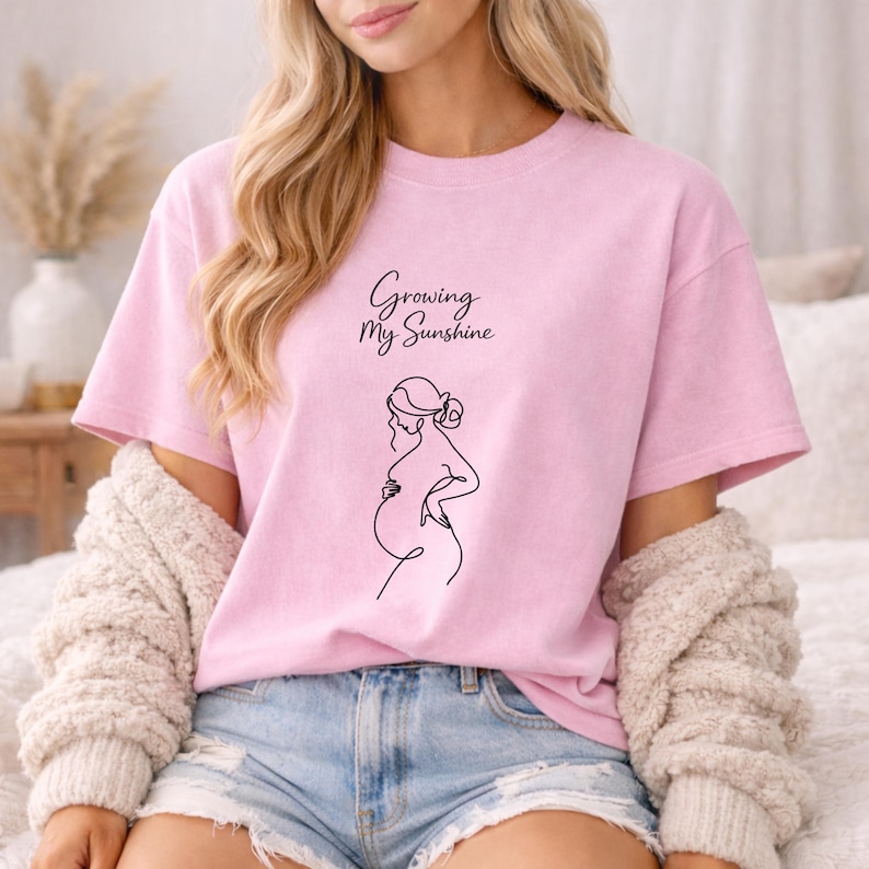 Puede incluir: Camiseta rosa claro con el texto "Growing My Sunshine" y un dibujo lineal negro de una persona embarazada. La camiseta tiene mangas cortas y un corte holgado.