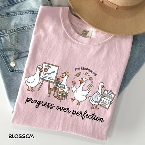 Op de afbeelding: Een lichtroze t-shirt met een afbeelding van vier witte ganzen die verschillende activiteiten uitvoeren, inclusief de tekst "GOAL: baseline progress", "SKILL BUILDER PLAN", "FUN REINFORCERS", "tokens reinforcer choice" en "progress over perfection".