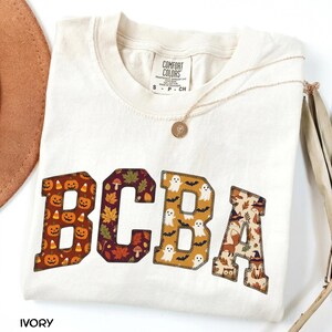 Könnte beinhalten: Ein elfenbeinfarbenes Kurzarm-T-Shirt mit Rundhalsausschnitt und einem Comfort Colors Markenetikett. Auf der Vorderseite befinden sich die Buchstaben BCBA in College-Schrift, jeweils gefüllt mit Mustern aus Kürbissen, Herbstblättern, Geistern und Füchsen.