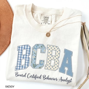 Op de afbeelding: Een ivoorkleurig t-shirt met de letters BCBA in een college-lettertype met diverse blauwe patronen, waaronder gingham, bloemen, stippen en strepen. Onder de letters staat de tekst Board Certified Behavior Analyst in een donkerblauw handschrift.