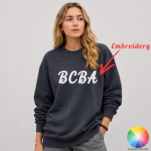Könnte beinhalten: Ein dunkel anthrazitfarbenes Sweatshirt mit Rundhalsausschnitt und dem weißen gestickten Text BCBA auf der Brust. Die Schrift ist in einer kursiven Schriftart gehalten. Ein roter Pfeil zeigt auf den Text mit dem Wort Embroidery in roter Schrift.