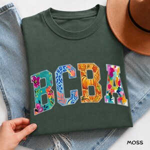 Könnte beinhalten: Ein dunkelgrünes T-Shirt mit den Buchstaben BCBA in einer College-Schriftart. Jeder Buchstabe hat ein eigenes Muster: Das B zeigt pinke und türkise Blumen, das C blaue und orangefarbene Wirbel, das B orangefarbene Kürbisse und das A bunte Farbspritzer.