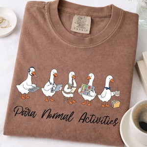 Puede incluir: Una camiseta marrón de cuello redondo con un gráfico de cinco gansos blancos con ropa de oficina realizando diversas tareas, con el texto "Para Normal Activities" impreso en letra cursiva negra debajo de la ilustración. La camiseta tiene una etiqueta Comfort Colors.