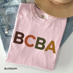 Könnte beinhalten: Ein hellrosa T-Shirt mit den Buchstaben BCBA in brauner, orangefarbener, olivgrüner und senfgelber Blockschrift. Das T-Shirt hat ein Comfort Colors-Etikett am inneren Halsausschnitt. Der Text Blossom ist in der unteren linken Ecke abgedruckt.