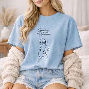 Puede incluir: Camiseta azul claro con el texto "Growing My Sunshine" y un dibujo lineal negro de una persona embarazada. La camiseta es de un material suave y tiene mangas cortas.