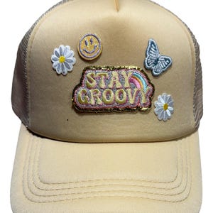 Könnte beinhalten: Beige Trucker-Cap mit Mesh-Rückseite und gebogenem Schirm. Verziert mit aufgestickten Patches, darunter ein Smiley, ein Schmetterling, Gänseblümchen und der Schriftzug "Stay Groovy" mit einem Regenbogen.