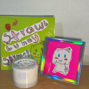 May include: A clear jar of white slime with colorful sprinkles, next to a green box with the words "Strawbunny Slime" and a drawing of a bunny. A pink framed drawing of a bunny is also present.