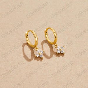 May include: Two gold-coloured hoop earrings, each with a small, flower-shaped charm. The charms are adorned with five clear, sparkling stones. The earrings are presented on a light brown surface.