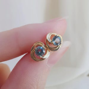 Peut inclure: Une paire de boucles d'oreilles à clous en or avec une pierre précieuse bleue ronde au centre, entourée d'une monture métallique circulaire en spirale. Le design présente de l'or poli qui s'enroule autour de la pierre en un effet tourbillon.