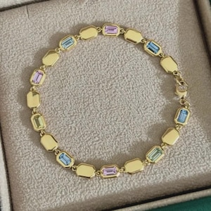 May include: A gold-toned chain bracelet featuring alternating rectangular links and bezel-set emerald-cut gemstones in pastel shades of pink, light blue, and pale green. The bracelet includes a secure clasp mechanism and a polished metallic finish.