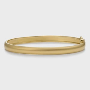 Może przedstawiać: Złota bransoletka typu bangle o gładkim, matowym wykończeniu. Okrągły element posiada mechanizm otwierania na zawiasie z jednej strony, co umożliwia bezpieczne otwieranie i zamykanie. Metal ma jednolity, delikatny żółty odcień na całej powierzchni.