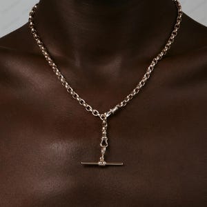 Puede incluir: Un collar plateado con un diseño de eslabones de cadena. El collar presenta un colgante con forma de barra en T. El collar se muestra sobre un fondo oscuro.