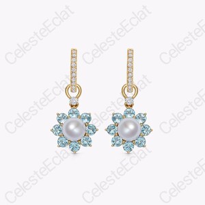 May include: A pair of gold drop earrings featuring a central white pearl surrounded by a halo of light blue round-cut gemstones. The earring posts are accented with small clear stones, and the design forms a flower-like shape with a dangling pendant.