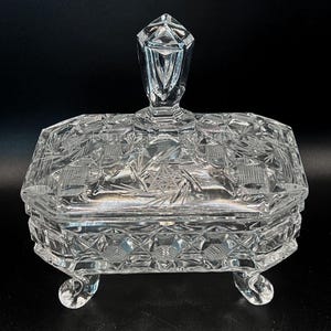 May include: A clear, rectangular crystal trinket box with a lid and four small feet. The box features intricate etched patterns, including geometric shapes and starburst designs. The lid has a faceted, diamond-shaped handle.