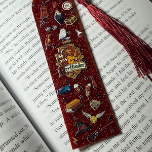 May include: A red glittery bookmark with various Harry Potter-themed charms, including a Gryffindor crest, a golden snitch, and a Hogwarts acceptance letter. The bookmark has a red tassel and is placed on an open book.