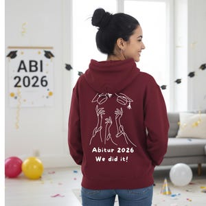 Puede incluir: Una sudadera con capucha granate con un gráfico blanco en la espalda que dice "Abitur 2026 We did it!" y un letrero a juego en el fondo. La sudadera con capucha tiene una capucha y la lleva una persona.