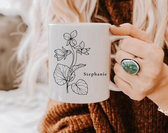 May include: White ceramic mug featuring a black floral illustration and the name "Stephanie". The mug has a curved handle and is held by a person. The design includes detailed leaves and flowers.