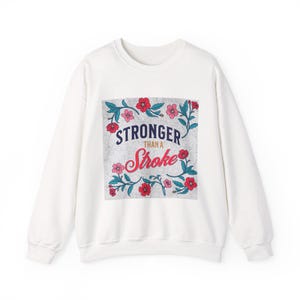 Può includere: Felpa bianca con la scritta "STRONGER than a Stroke" in blu navy e rosso, circondata da un motivo floreale con fiori rossi e rosa e foglie verdi. Girocollo.