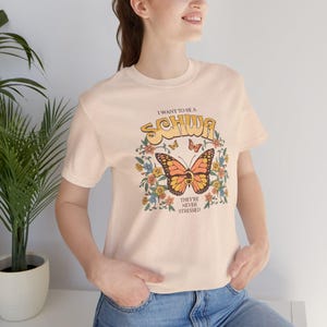 May include: A light peach-colored t-shirt with a butterfly graphic. The graphic includes the text "I want to be a Schwa, They're Never Stressed" in a retro font, surrounded by flowers and butterflies. The shirt is short-sleeved.