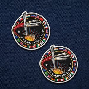 May include: Two embroidered patches with a circular design. The patches feature a spacecraft, a rising sun, and the text "PROJECT HAIL MARY." The design is framed by a ring of international flags. The patches are on a dark blue fabric background.