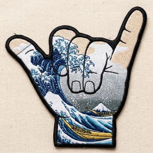 May include: Embroidered patch in the shape of a hand making the shaka sign. The design inside features a depiction of Hokusai's "The Great Wave off Kanagawa" with blue waves, a mountain, and boats.