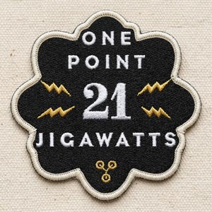 May include: Black embroidered patch with a scalloped edge. The patch features the text "ONE POINT 21 JIGAWATTS" in white, with two lightning bolt designs in gold. A small gold design is at the bottom.