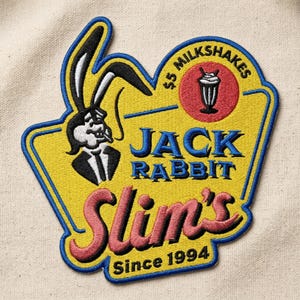 May include: Embroidered patch featuring a cartoon rabbit in a suit, smoking a cigarette, with the text "Jack Rabbit Slim's" in pink and blue. The patch also includes a milkshake graphic and the text "Since 1994".