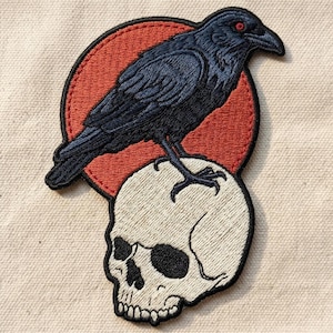 May include: Embroidered patch featuring a black raven with red eyes perched atop a white skull. The raven is set against a red circle, with black stitching outlining the design. The patch is on a beige fabric background.