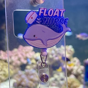May include: A badge reel featuring a cartoon whale with a nurse's cap and the words "FLOAT NURSE" in pink and blue glitter. The reel has a clear plastic strap and a metal clip for attaching to clothing.