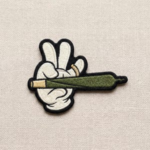 May include: Embroidered patch featuring a cartoon hand making a peace sign, holding a green and tan rolled item. The hand is white with black outlines, and the rolled item is green with a tan tip. The patch is on a beige fabric background.