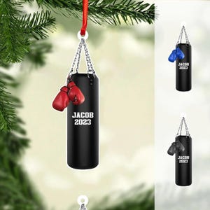 May include: A black punching bag Christmas ornament with red boxing gloves. The ornament is suspended by a silver chain and a red ribbon. The text "JACOB 2023" is printed on the bag.